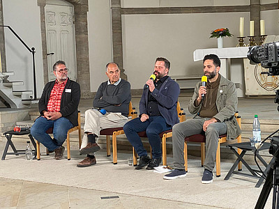 Vier Männer sitzen im Halbkreis auf Stühlen. Die beiden rechten halten gelbe Mikofone in der Hand. Es sind die Teilnehmenden der Podiumsdiskussion "Combattants for Peace", die am 23.04.2026 in Oldenburg stattfand.en. 